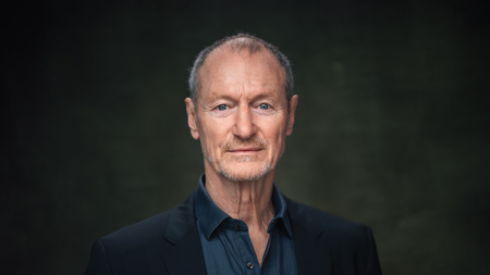 Porträtt av äldre man med kort hår och skägg, klädd i mörk kavaj och skjorta mot mörk bakgrund.