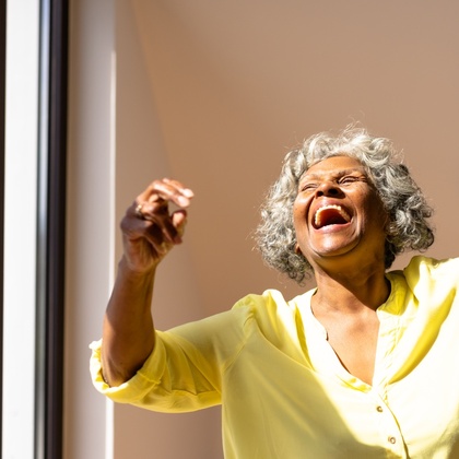 Äldre kvinna i gul skjorta skrattar och dansar med en man i ett soligt rum.