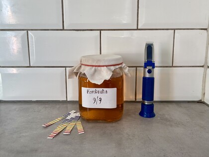 En burk kombucha med tyglock, pH-stickor och ett blått refraktometer på en köksbänk.