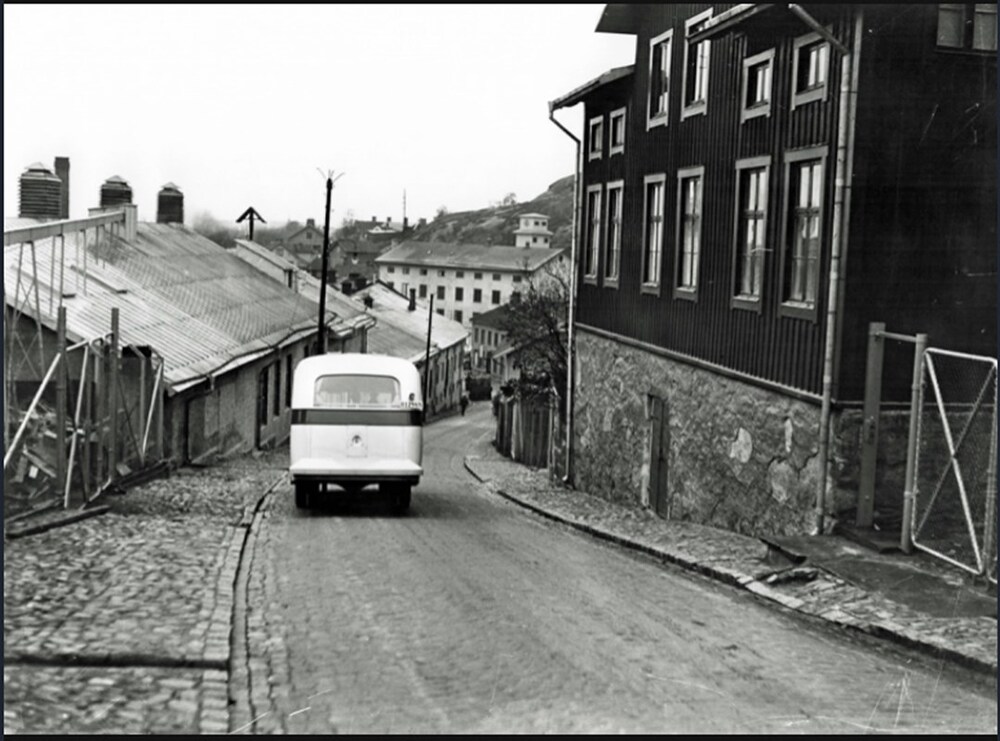 En svartvit bild av en gammal bil som kör nerför en kullerstensgata mellan äldre hus.
