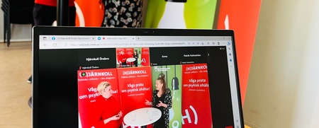 Två personer samtalar vid ett ståbord framför röda rollups, med en laptop i förgrunden som visar samma scen på skärmen.