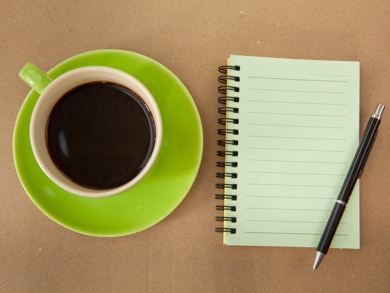 En kopp svart kaffe på ett grönt fat bredvid ett tomt spiralblock och en penna på ett brunt bord.