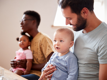 Två män sitter vid ett bord, var och en håller ett litet barn i knät, i ett ljust rum.