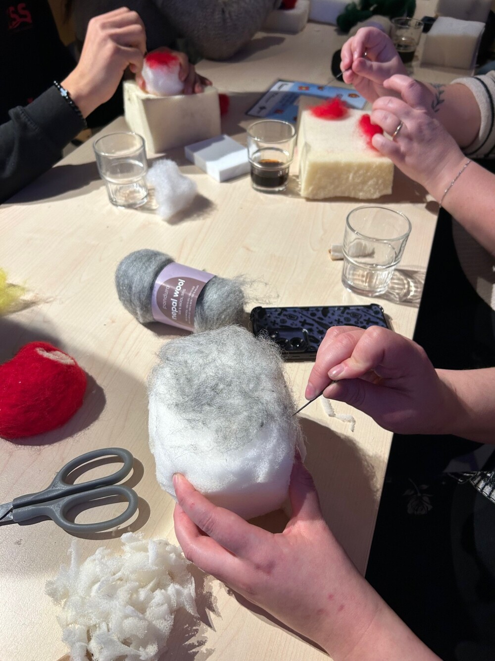 Personer arbetar med tovning av ull på ett bord med verktyg, glas och material utspridda.