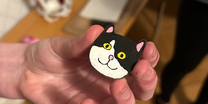 En hand håller en liten figur av en svartvit katt med gula ögon, i ett rum med trägolv och strykjärn i bakgrunden.