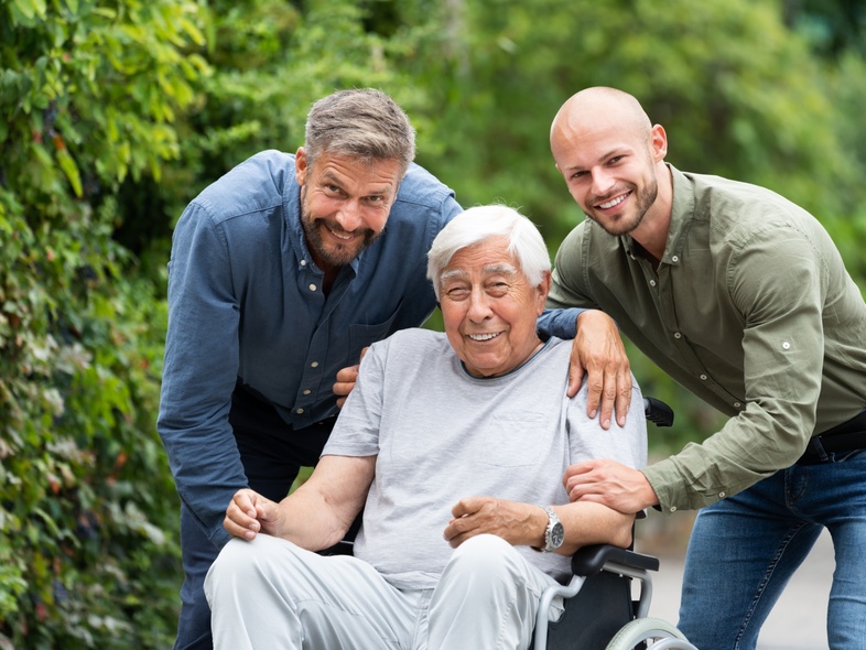 Två yngre män står bredvid en äldre man i rullstol utomhus, alla ler mot kameran.