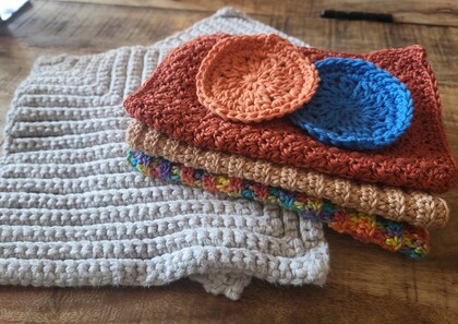 Flera virkade grytlappar och runda underlägg i olika färger ligger staplade på ett träbord.