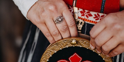 Närbild av händer som håller en traditionell väska med röd brodyr och mässingsdetaljer, klädd i folkdräkt.