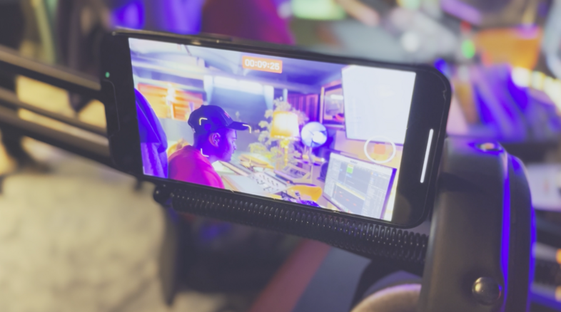 En smartphone filmar en person vid en ljudstudio med flera skärmar och utrustning.
