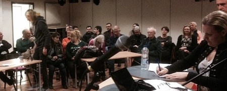 Ett möte eller seminarium med människor som sitter i ett rum, några vid ett bord med mikrofon och papper.