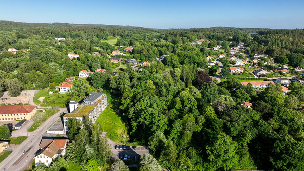 Flygfoto över en grönskande småstad med hus, vägar och tät skog under en klarblå himmel.