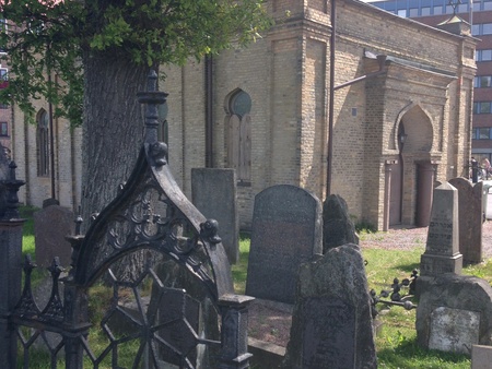 Gammal judisk kyrkogård med gravstenar och en synagoga i bakgrunden, omgiven av träd och gräs.