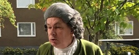 Man i grön 1700-talsdräkt står utomhus vid ett bord med vit duk och tårta.