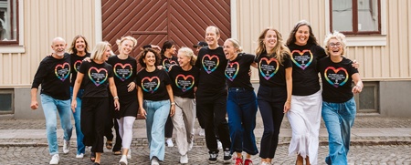 En grupp människor går tillsammans på en kullerstensgata framför en träbyggnad, alla bär svarta t-shirts med regnbågshjärtan.