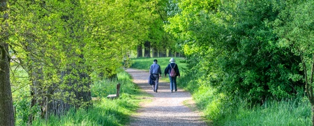 Två personer promenerar på en grusväg omgiven av grönskande träd och buskar en solig dag.