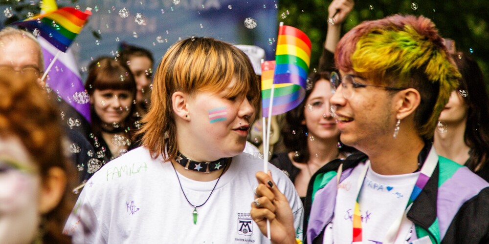 Två personer håller regnbågsflaggor på en prideparad, omgivna av bubblor och andra deltagare.