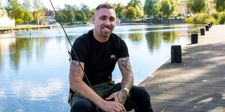 En man sitter på en brygga vid en sjö och fiskar, omgiven av träd och vatten.