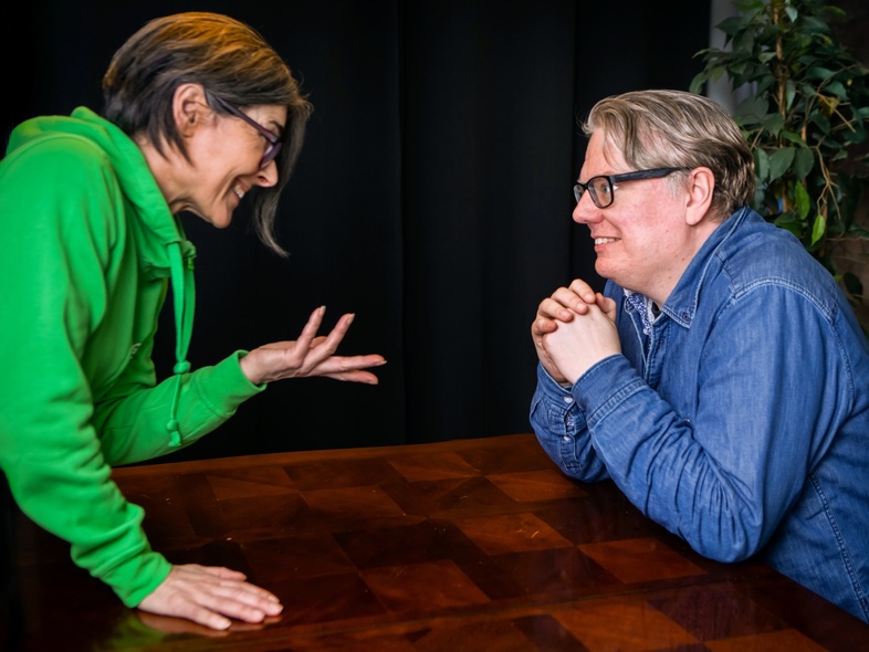 Två personer samtalar vid ett träbord, en i grön tröja gestikulerar, den andra i blå skjorta lyssnar leende.