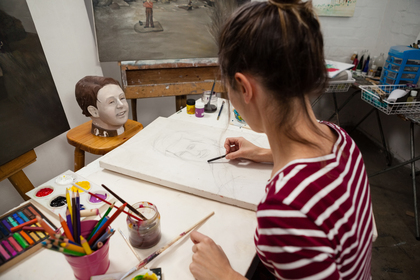 En kvinna tecknar ett porträtt vid ett konstbord med färgpennor och penslar i en ateljé.