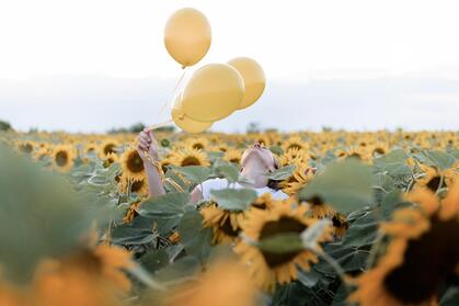 En person håller tre gula ballonger i ett fält av solrosor under en ljus himmel.