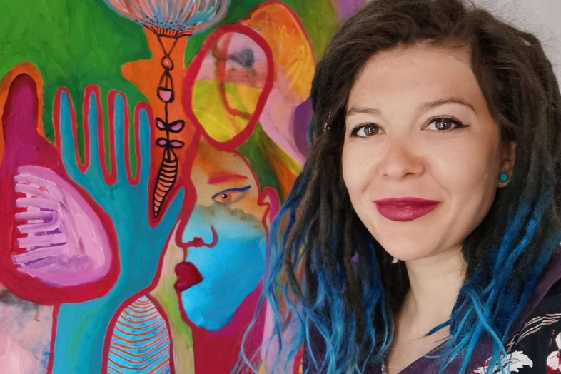 Kvinna med blått hår står bredvid en färgstark abstrakt målning på väggen.