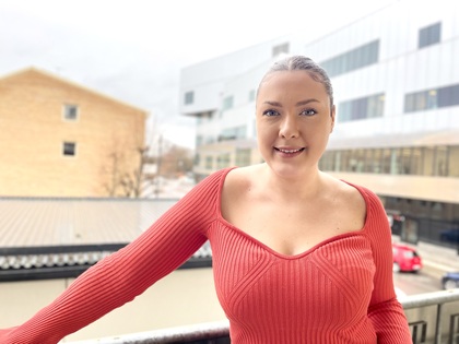 Kvinna i röd tröja står på en balkong med moderna byggnader i bakgrunden.