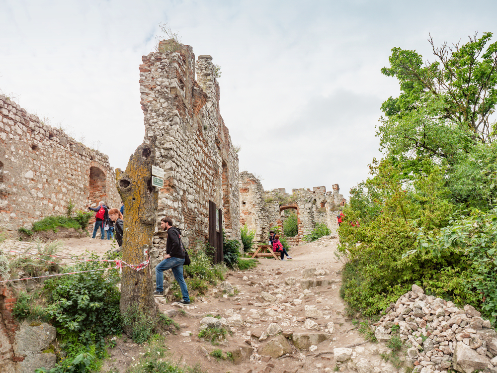 Ruiner av en gammal stenbyggnad med flera personer som promenerar bland stenarna och grönska.