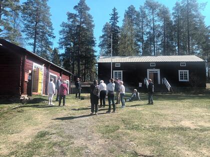 En grupp människor står utomhus mellan två gamla trähus omgivna av höga tallar en solig dag.