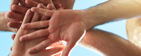 Flera personer står i en cirkel och lägger sina händer ovanpå varandra mot en blå himmel.