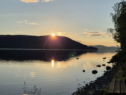 Solnedgång över en sjö med spegelblankt vatten, berg i bakgrunden och stenar längs strandkanten.