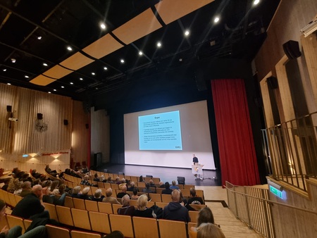 Föreläsning i en biosalong med publik, en person står vid scenen och visar en presentation på stor skärm.