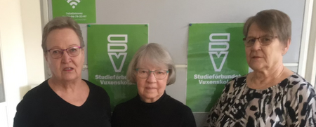 Tre äldre kvinnor står framför gröna skyltar med texten 'Studieförbundet Vuxenskolan'.