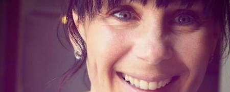 Leende kvinna med mörkt hår håller en vit kopp och tittar in i kameran.