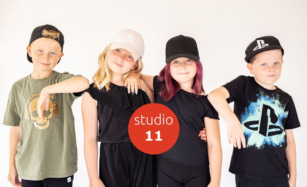 Fyra barn i kepsar poserar tillsammans framför en vit bakgrund med texten 'studio 11' i mitten.