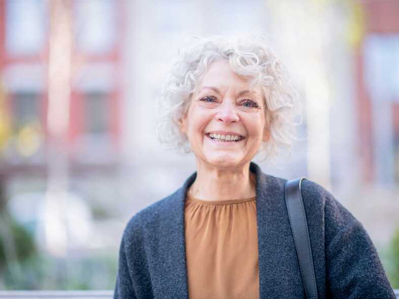 En äldre kvinna med lockigt hår ler mot kameran. Hon är klädd i blå kavaj och har en väska över axeln. 