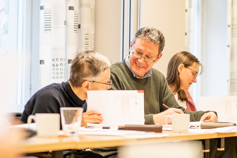 Tre personer sitter vid ett bord i ett ljust rum och samtalar, med papper och kaffekoppar framför sig.