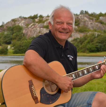 Person spelar akustisk gitarr utomhus vid en sjö med berg i bakgrunden.