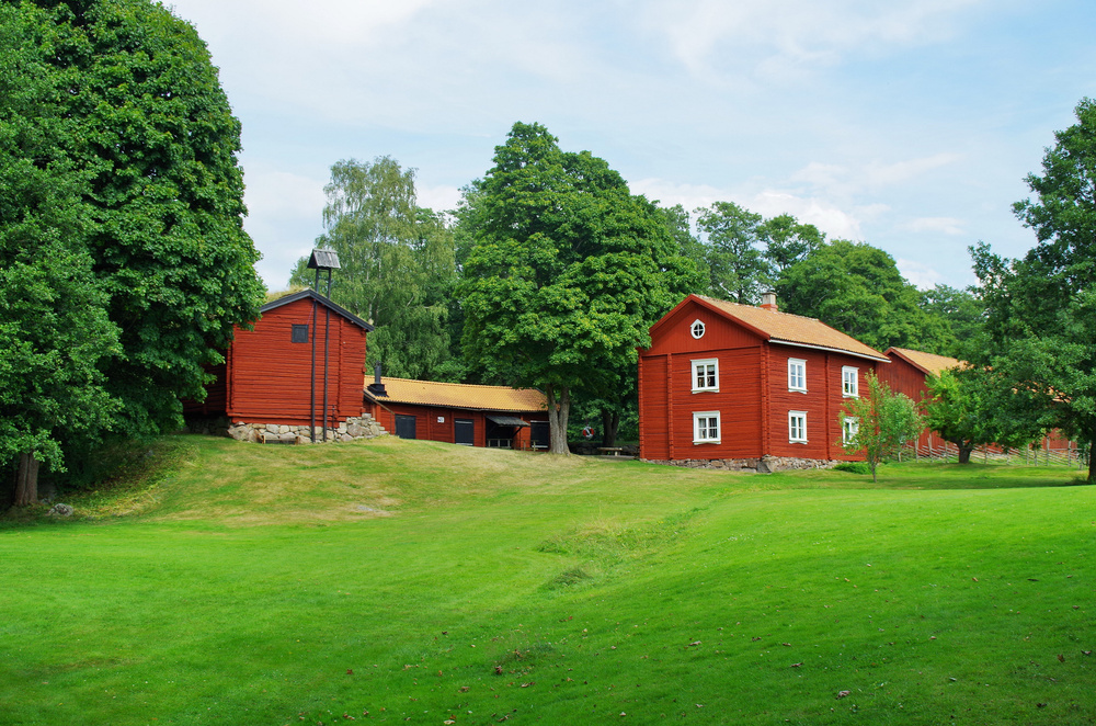 Röda trähus omgivna av grönskande träd och gräsmatta under blå himmel.