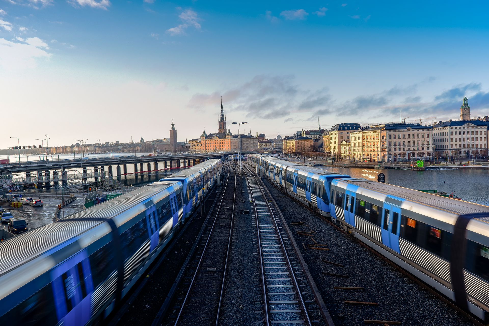 Två tunnelbanetåg kör på parallella spår genom centrala Stockholm med stadens siluett i bakgrunden.