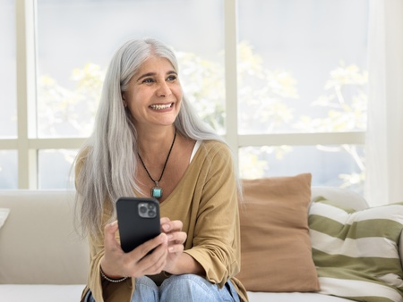 Kvinna med långt grått hår sitter i soffa och håller en mobiltelefon i vardagsrum med stora fönster.
