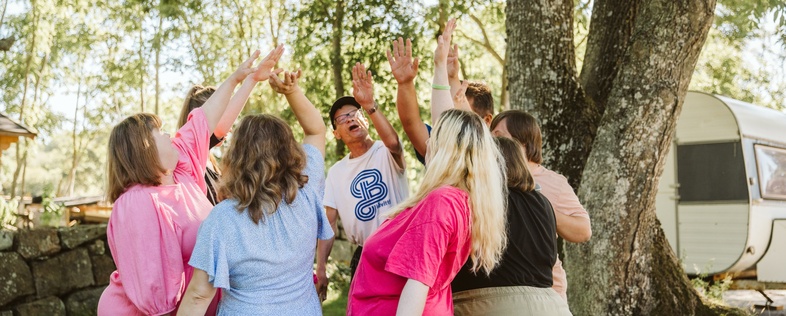 En grupp människor står i en ring utomhus och sträcker upp händerna mot mitten under ett träd.
