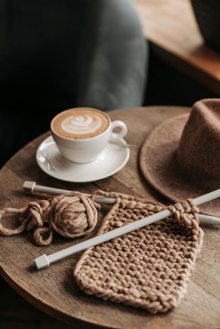 En kopp cappuccino, ett brunt garnnystan med stickning och en brun hatt på ett träbord.
