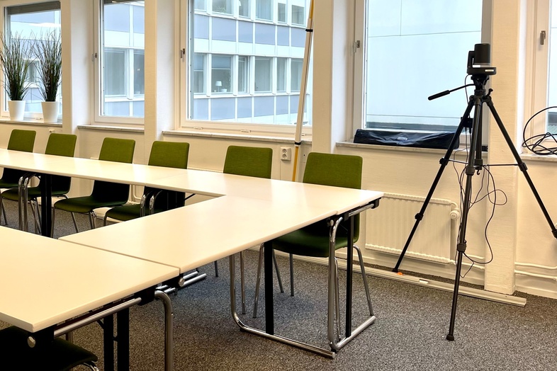 Ljust konferensrum med gröna stolar, vita bord, stora fönster och en kamera på stativ.