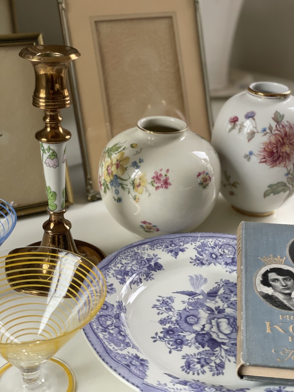 Stilleben med porslinsvaser, blåvit tallrik, glas, ljusstake och bok på ett bord.
