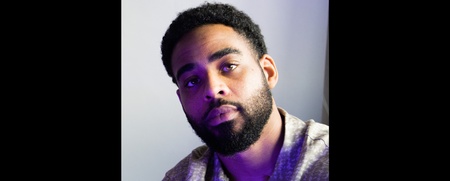 Man med kort lockigt hår och skägg sitter mot en ljus bakgrund, bär skjorta och halsband.