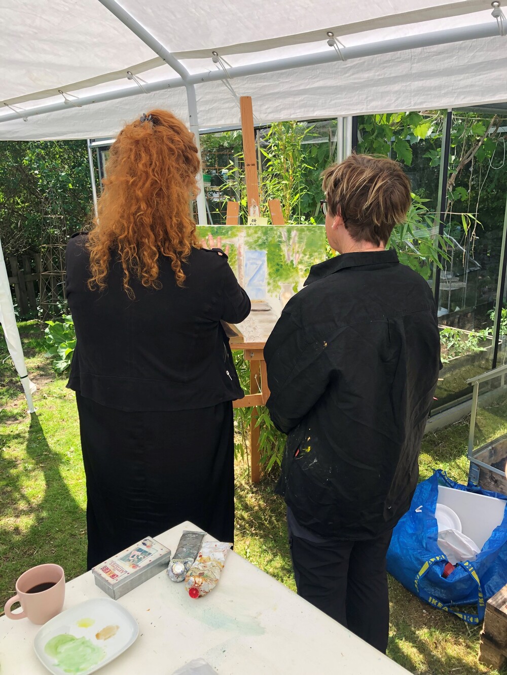 Två personer står under ett tält och målar vid ett staffli i en trädgård.