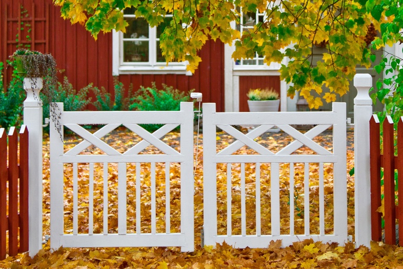 Vit trägrind framför ett rött hus, omgiven av höstlöv och gröna trädgrenar.