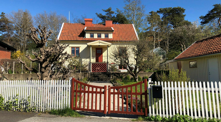 Ett gult trähus med rött tegeltak, vit staket och trädgård i soligt väder.