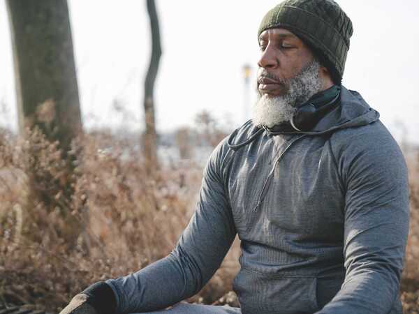 En äldre man med skägg sitter utomhus och mediterar. Han har en grå hoodie och mössa på sig.