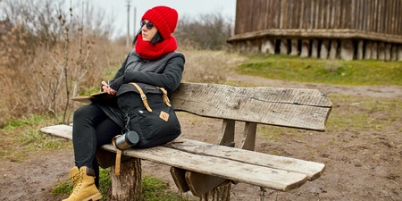 Person med röd mössa och halsduk sitter på en träbänk utomhus och skriver i ett block.